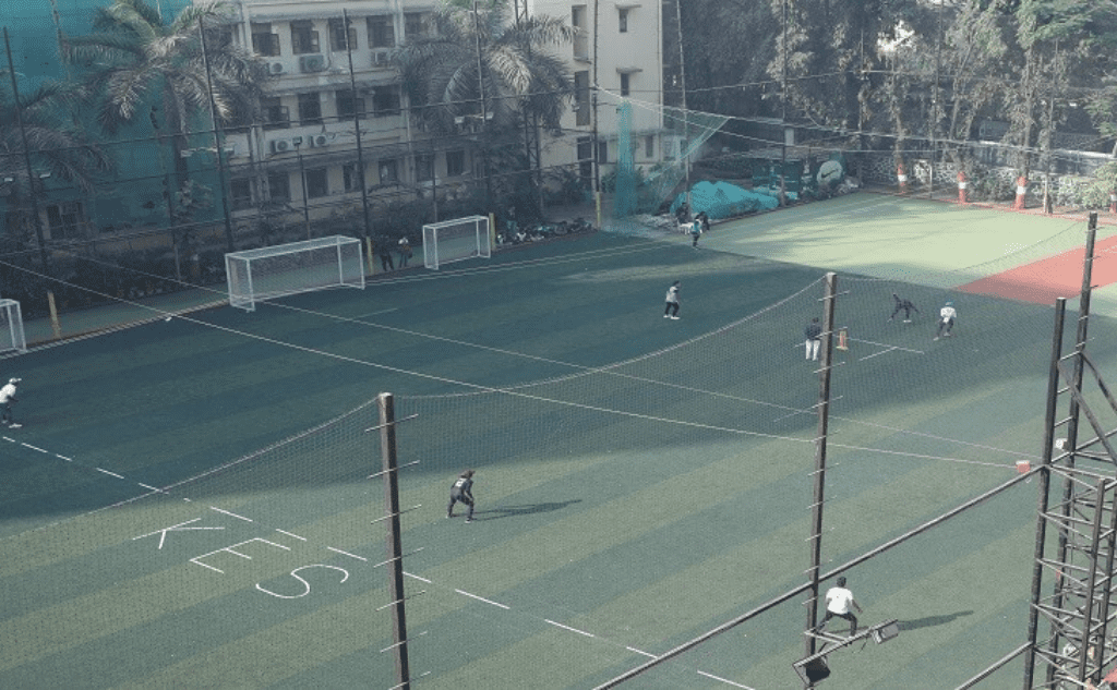 Outdoor fenced soccer field with multiple goals and players warming up on a green artificial turf, buildings in background.